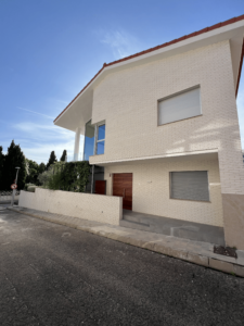 Imagen de una casa moderna de dos pisos con una fachada de ladrillo claro y un techo inclinado con tejas rojas. La entrada principal tiene una puerta de madera oscura, y al lado se encuentra una ventana con persiana metálica cerrada. Un muro bajo de ladrillo delimita la propiedad, y en el fondo se observan árboles y vegetación.