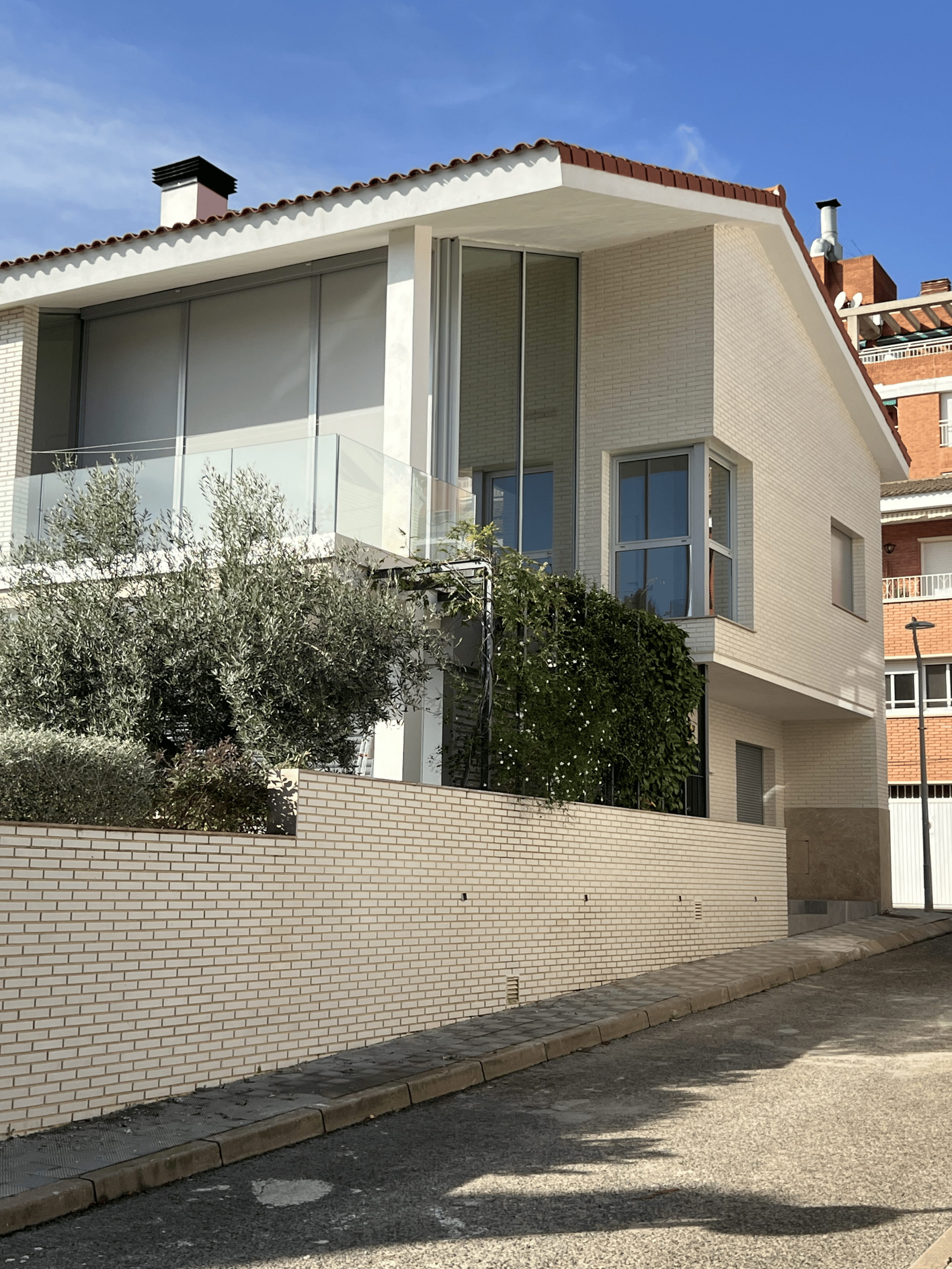 Imagen de una casa moderna de dos pisos con una fachada de ladrillo claro y un techo inclinado con tejas rojas. La vivienda cuenta con amplios ventanales, una terraza con barandilla de cristal y vegetación en la entrada. Un muro de ladrillo rodea la propiedad, y en el fondo se observan otros edificios residenciales de estilo más tradicional.