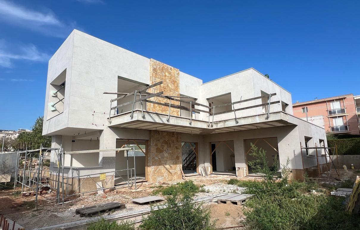 Imagen de una casa de dos pisos en construcción con un diseño moderno y minimalista. La estructura cuenta con amplias terrazas, ventanales grandes y una fachada de color claro con detalles en piedra. Hay andamios y materiales de construcción en el sitio, mientras que en el fondo se observan otros edificios residenciales.